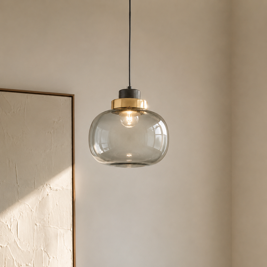 A modern pendant light with a clear glass dome shade, a brushed brass metal collar, and a black ceiling mount, hanging in a minimalist room with a textured beige wall.