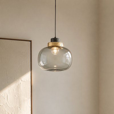 A modern pendant light with a clear glass dome shade, a brushed brass metal collar, and a black ceiling mount, hanging in a minimalist room with a textured beige wall.