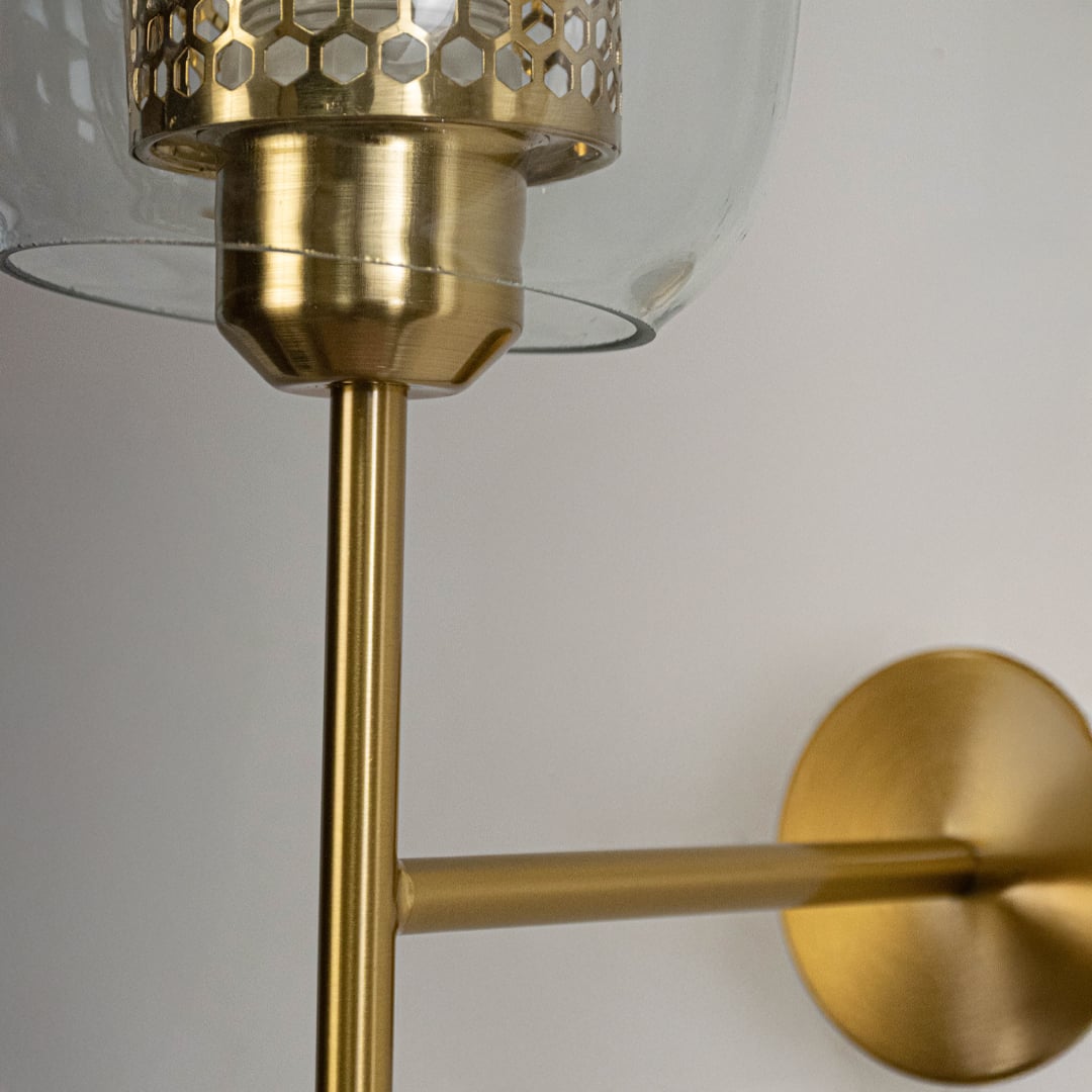 Closeup view of wall light showing clear glass shade with honeycomb metal inner cylinder and brushed gold metal arm mounted on a white wall.