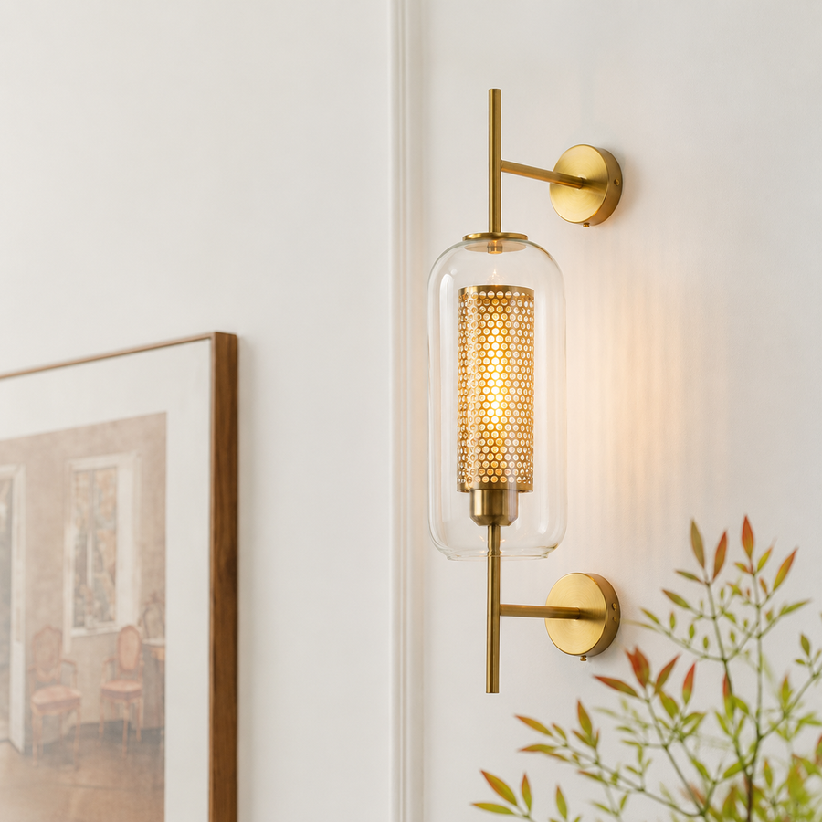 A modern wall sconce light fixture with a gold-finished metal frame, a clear cylindrical glass shade, and a glowing warm bulb inside, mounted on a white wall next to a framed picture and green leaves.