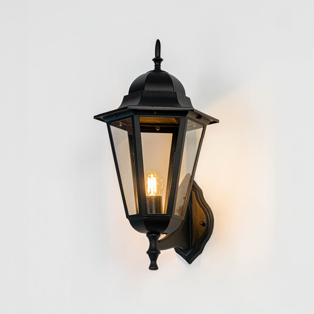 Black metal and clear glass outdoor wall lantern mounted on exterior wall creating warm entryway lighting ambiance shown Light on white BG.