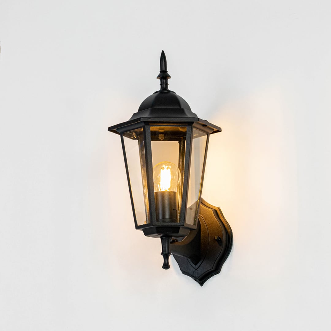 Illuminated outdoor wall lantern on white background highlighting black finish clear glass and soft golden glow.