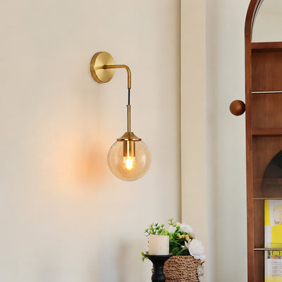 Mid century gold brass wall sconce with amber glass globe light installed in a cozy bedroom corner near a wooden wardrobe.