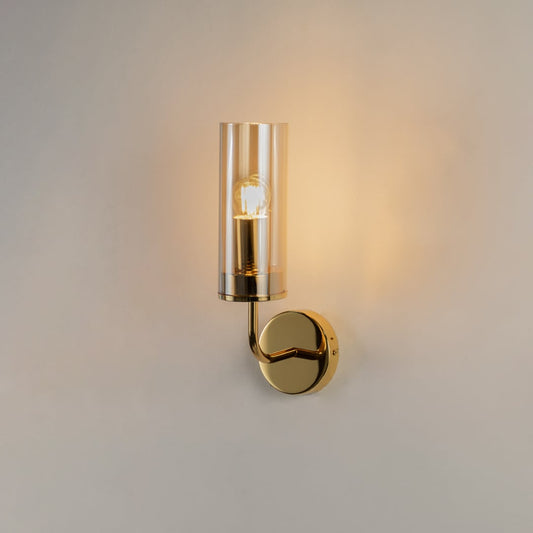 Illuminated gold wall lamp with clear glass cylinder shade isolated on white background highlighting bright warm lighting for interiors.