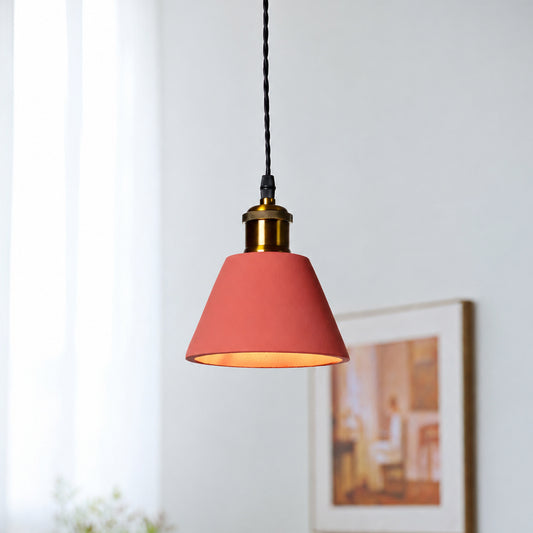 A modern pendant light with a terracotta pink cone-shaped shade, a brass metal collar, and a black hanging cord, shown in a softly lit room with a blurred framed picture in the background.