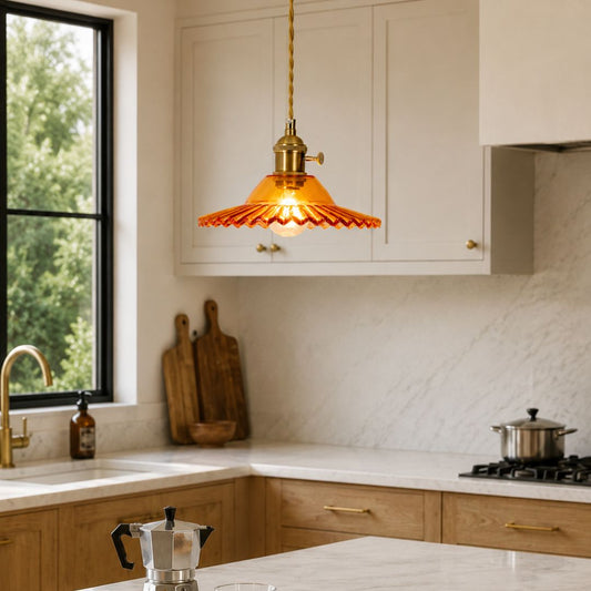 A warm amber glass pendant light with a scalloped edge hangs over a modern kitchen with white cabinets, a marble backsplash, and wood countertops.