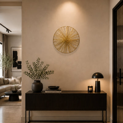 A modern styled interior featuring a round gold wire starburst wall art piece hanging on a beige wall above a dark wood console table with a black lamp and vase.