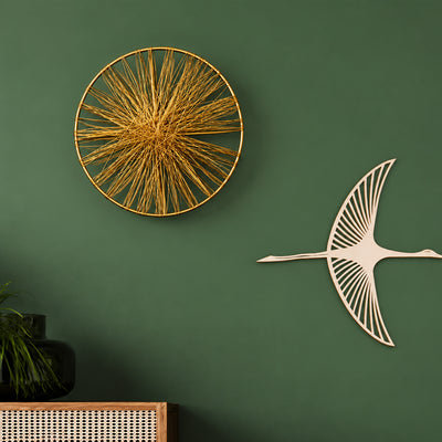Decorative wall art displayed on a dark green wall, featuring a circular gold wireburst sculpture and a white wooden crane silhouette mounted to the right. A small potted plant and woven furniture are partially visible at the bottom left.