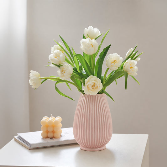 A pink ribbed ceramic vase filled with white tulips, placed on a white table next to a small yellow candle on a book against a soft beige wall.