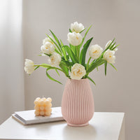 A pink ribbed ceramic vase filled with white tulips, placed on a white table next to a small yellow candle on a book against a soft beige wall.