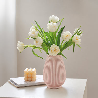 A pink ribbed ceramic vase filled with white tulips, placed on a white table next to a small yellow candle on a book against a soft beige wall.
