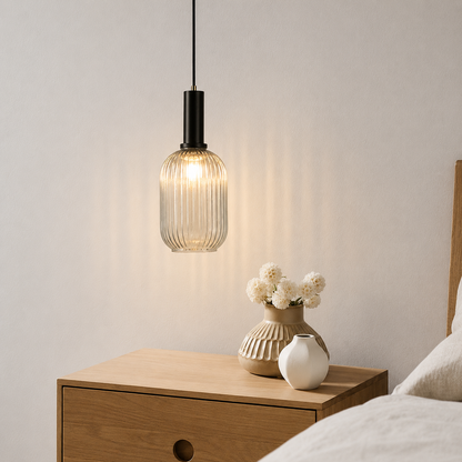 A modern pendant light with a ribbed clear glass shade and a black metal neck hangs above a wooden bedside table in a bedroom setting, with vases and bedding visible.