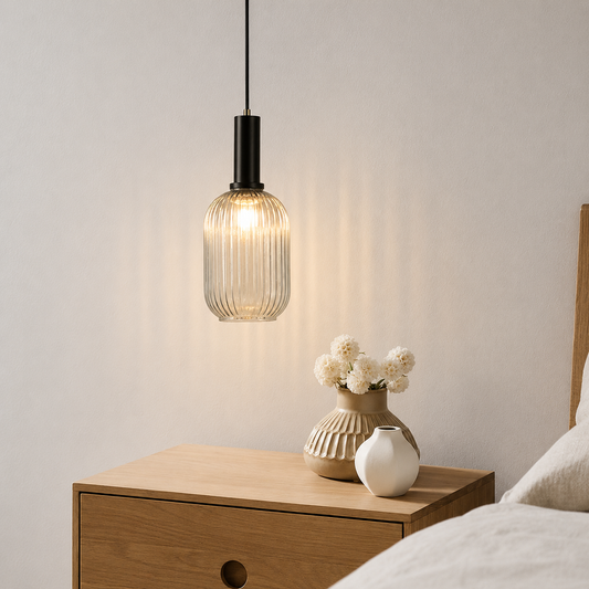 A modern pendant light with a ribbed clear glass shade and a black metal neck hangs above a wooden bedside table in a bedroom setting, with vases and bedding visible.