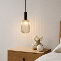 A modern pendant light with a ribbed clear glass shade and a black metal neck hangs above a wooden bedside table in a bedroom setting, with vases and bedding visible.