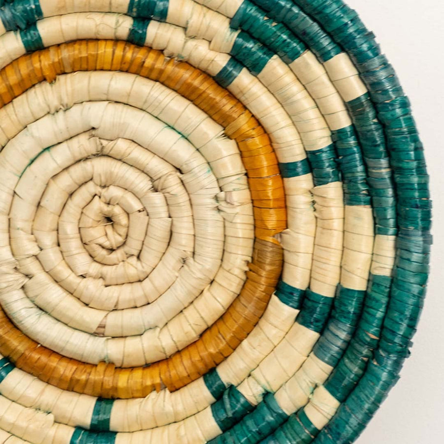A closeup view of the teal mustard and natural rattan wall decor plate showing its detailed sunburst inspired weave and textured handcrafted pattern for vibrant decor styling.