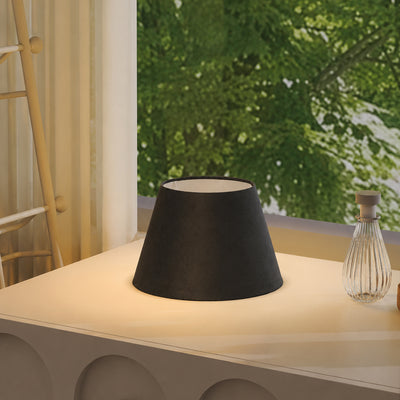 A black fabric lampshade with a warm white interior, glowing with a soft warm light, set on a white table in a modern room with a window showing green foliage outside.