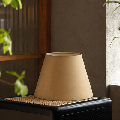 A beige fabric lampshade with a tapered conical shape, displayed on a black side table with a woven cane top, set against a textured brown wall with soft natural light.