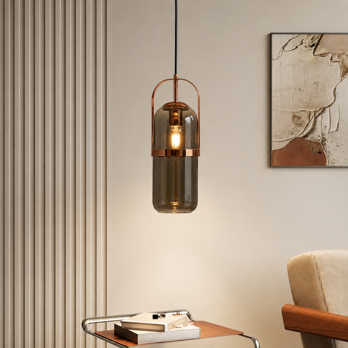 A modern cylindrical pendant light with a smoky translucent glass shade and copper-toned metal accents, hanging in a styled interior setting with a cream wall, framed art, and a side table.