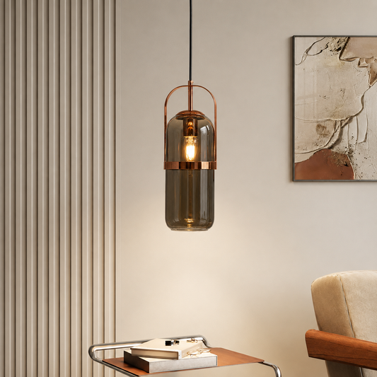 A modern cylindrical pendant light with a smoky translucent glass shade and copper-toned metal accents, hanging in a styled interior setting with a cream wall, framed art, and a side table.