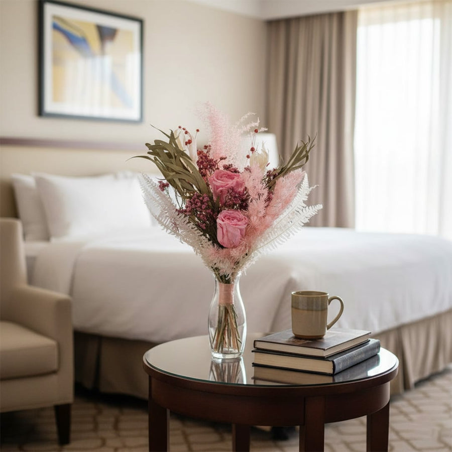 Main lifestyle image of preserved pink floral bouquet arranged in glass vase on bedside table creating elegant hotel bedroom decor ambience.