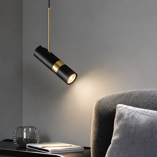 A modern black cylindrical pendant light with a gold band and gold hanging rod, shown lit in a styled interior next to a gray upholstered chair.