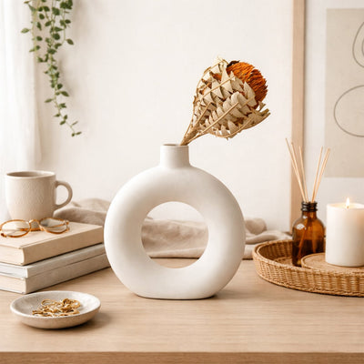 Orange dried banksia flower stem in a white donut ceramic vase on a wooden table next to books and a lit candle.