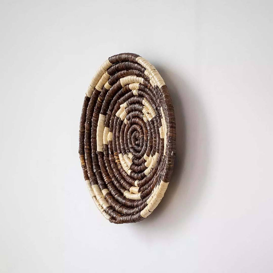 Handwoven seagrass round wall basket decor in natural beige and dark brown with raised spiral woven profile on white.