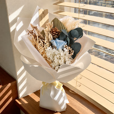 A dried flower bouquet wrapped in white paper and tied with a pale yellow ribbon, placed on a wooden windowsill with sunlight and window blinds in the background.