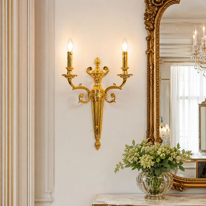An ornate gold wall sconce with two lit candle-style bulbs mounted on a cream-colored wall in a luxurious interior setting with a large gold mirror and a glass vase of flowers.