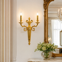 An ornate gold wall sconce with two lit candle-style bulbs mounted on a cream-colored wall in a luxurious interior setting with a large gold mirror and a glass vase of flowers.