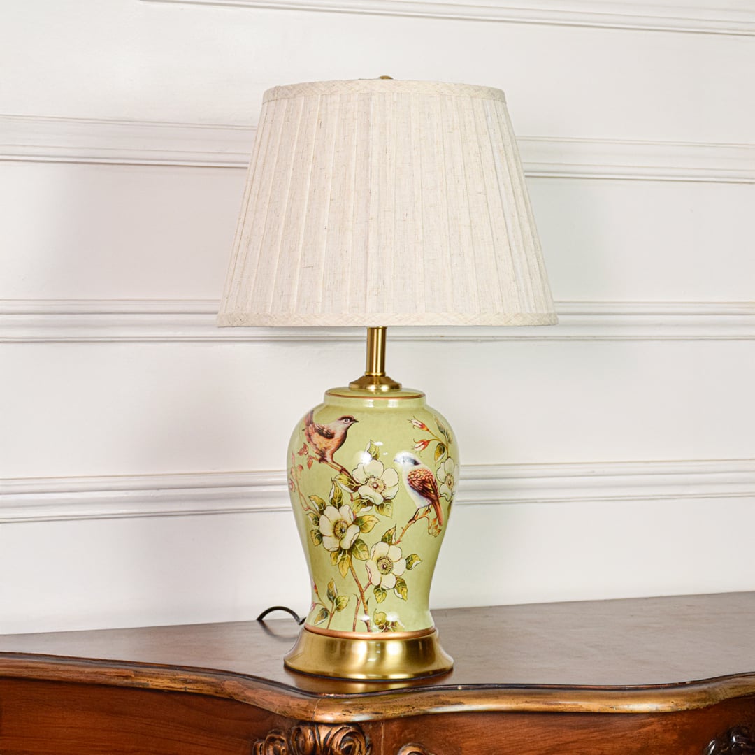 Decorative green ceramic table lamp with bird and floral design brass base and fabric shade on soft off white background.
