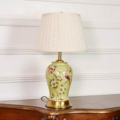 Decorative green ceramic table lamp with bird and floral design brass base and fabric shade on soft off white background.