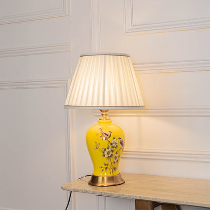 Table lamp glowing softly with yellow ceramic base and white pleated shade on a white background for warm decor styling.
