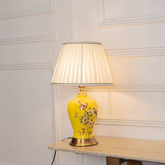 Table lamp glowing softly with yellow ceramic base and white pleated shade on a white background for warm decor styling.