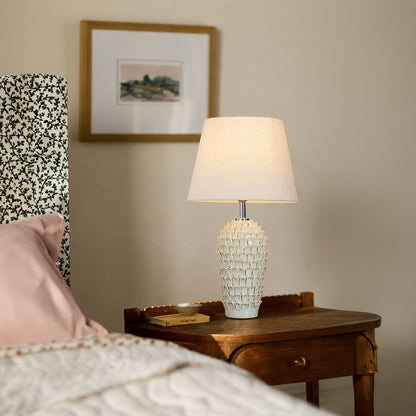 White textured ceramic table lamp with fabric shade placed on a bedside table creating warm ambient bedroom lighting.