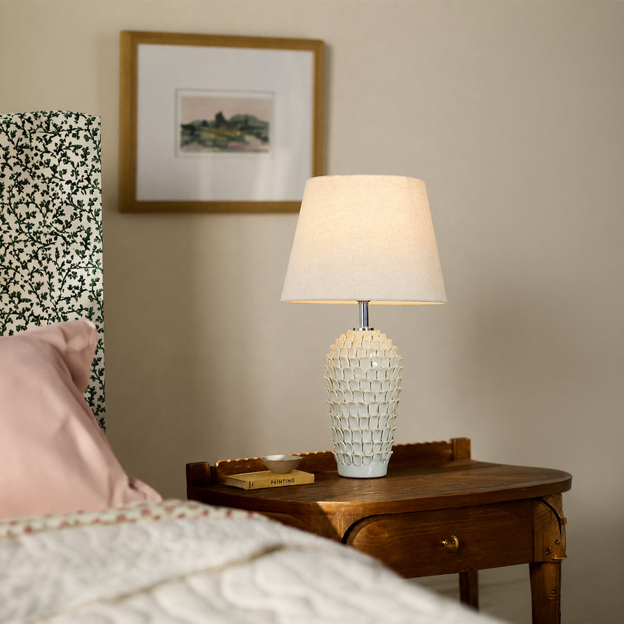 White textured ceramic table lamp with fabric shade placed on a bedside table creating warm ambient bedroom lighting.
