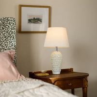 White textured ceramic table lamp with fabric shade placed on a bedside table creating warm ambient bedroom lighting.