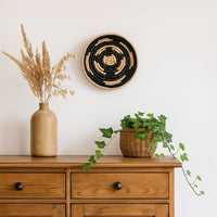 Lifestyle image showcasing a handcrafted round woven wall basket mounted on a white wall styled above a wooden console with plants and dried grass creating a calm modern home decor setting.