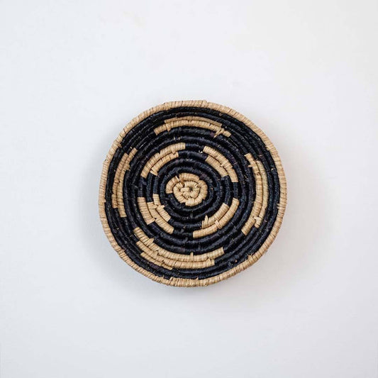 Handwoven seagrass round wall basket decor in natural beige and black with abstract spiral woven pattern on white background.