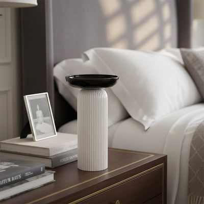 Lifestyle image of a minimalist cream pillar vase with a black dish top styled on a luxury bedroom nightstand table.