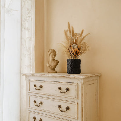 Small round black textured ceramic vase styled with dried pampas and protea flowers placed on a vintage wooden dresser.