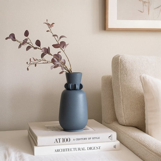 Modern blue ceramic decorative vase placed on stacked books beside sofa holding dried floral stems for elegant minimalist living room decor.