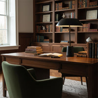 A black metal desk lamp glowing softly on a wooden study table creating a warm reading ambience in a classic home office setting.
