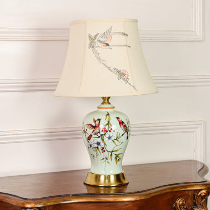 Decorative mint green ceramic table lamp with bird floral design gold metal base and fabric shade on off white backdrop.