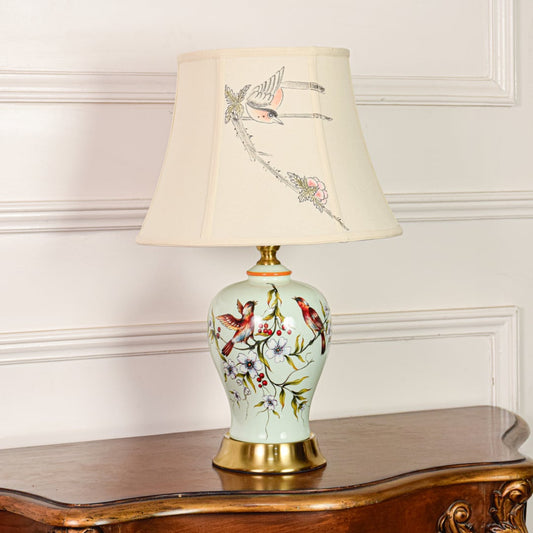 Decorative mint green ceramic table lamp with bird floral design gold metal base and fabric shade on off white backdrop.