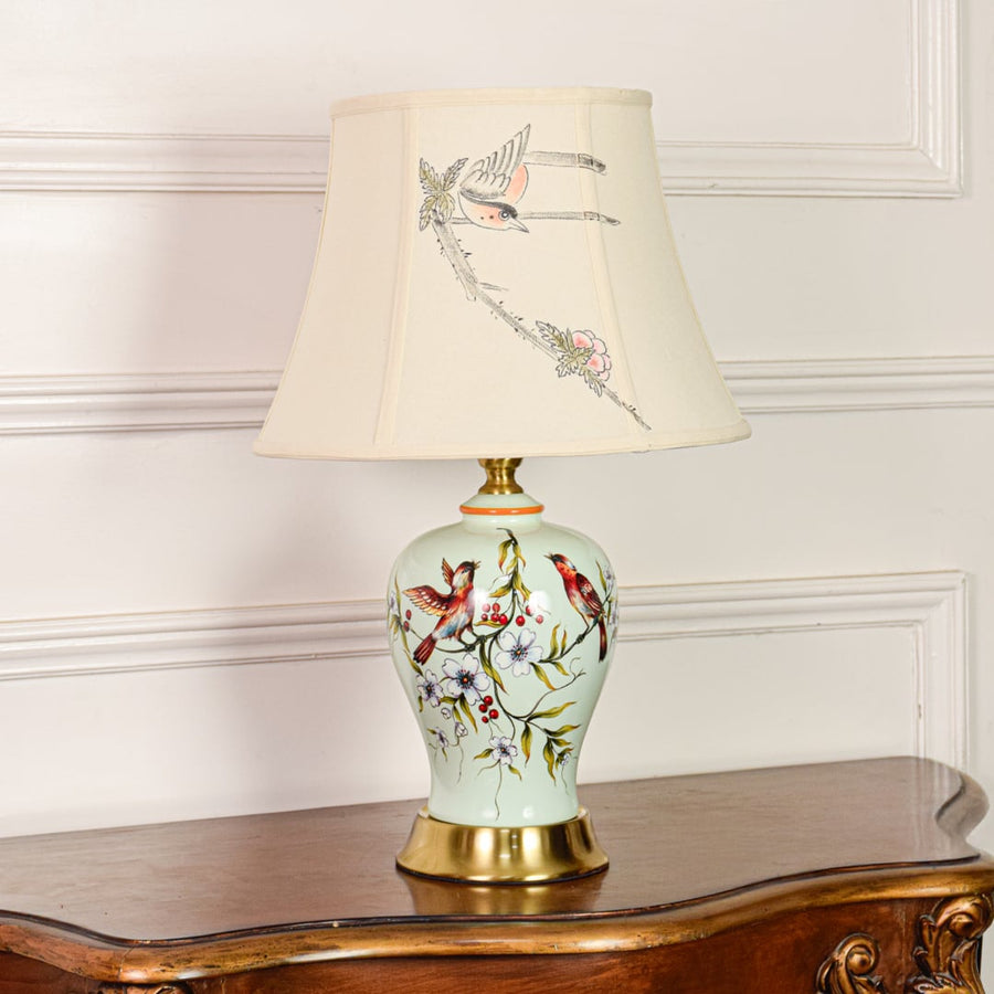 Decorative mint green ceramic table lamp with bird floral design gold metal base and fabric shade on off white backdrop.