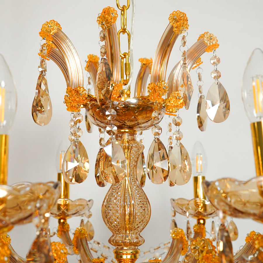 A close-up of a decorative chandelier featuring translucent amber glass arms, gold-toned metal hardware, and hanging crystal teardrop pendants with visible light bulbs.