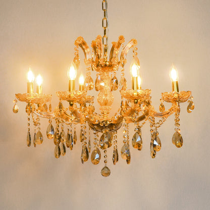 An ornate gold chandelier with six lit candle-style bulbs and hanging clear crystal teardrop pendants, shown against a plain light beige wall.