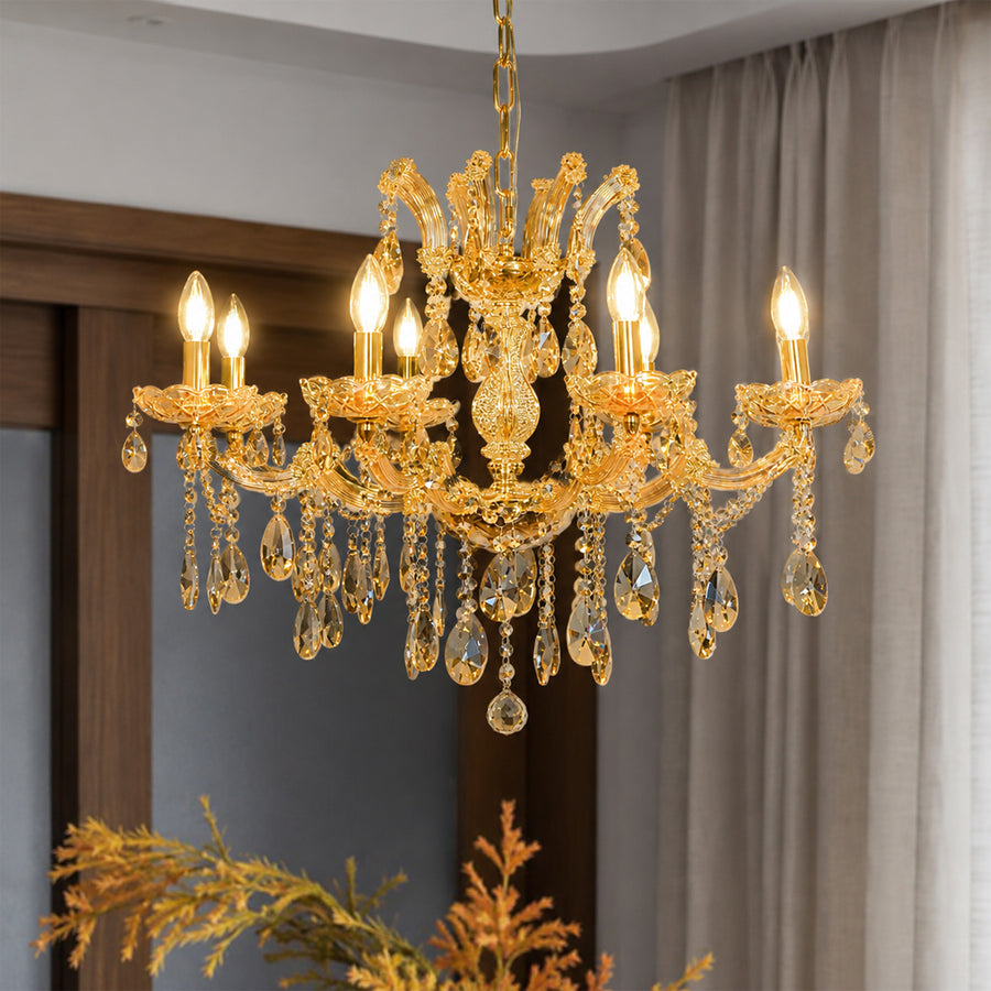 An ornate gold chandelier with clear crystal droplets and six lit candle-style bulbs, hanging in a room with beige curtains and a wooden door frame.