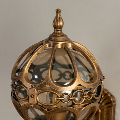 Closeup of gold metal outdoor wall lantern with dome glass design and ornate frame detailing for stylish decorative exterior lighting.
